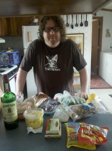 Ted and the Provisions A man with long curly hair, a beard and glasses standing behind a counter full of food items.